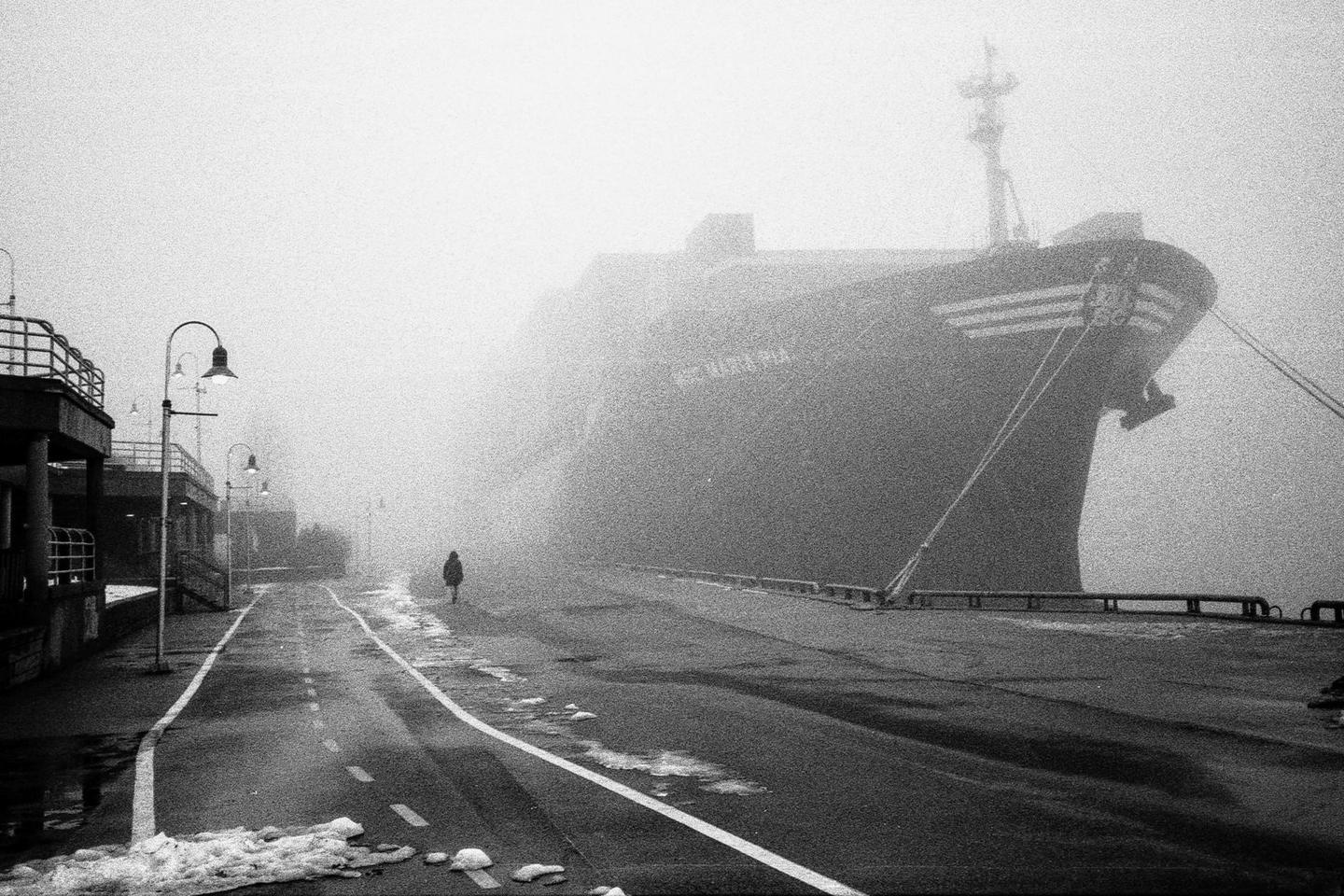 A person walks along a foggy street next to a large ship docked at a pier.