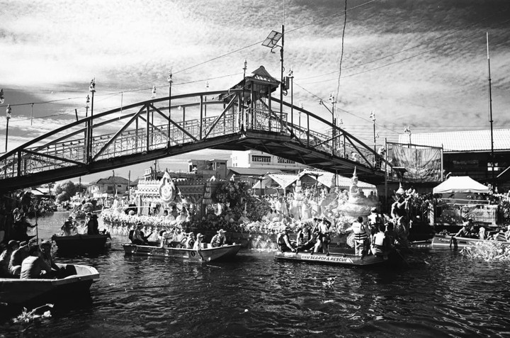 A black and white image shows a bridge over a canal with boats and people, with ornate decorations on the canal banks.