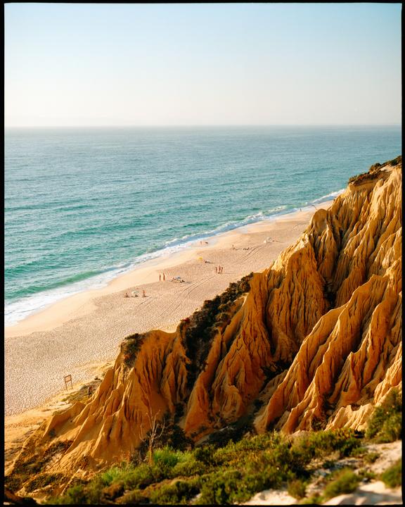 A sandy beach with people and umbrellas is visible below a cliff with eroded rock formations.