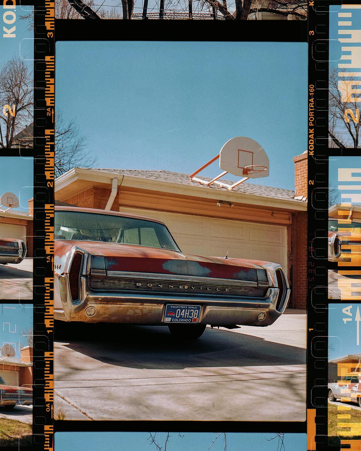A rusty red car is parked in a driveway in front of a garage with a basketball hoop on the roof.
