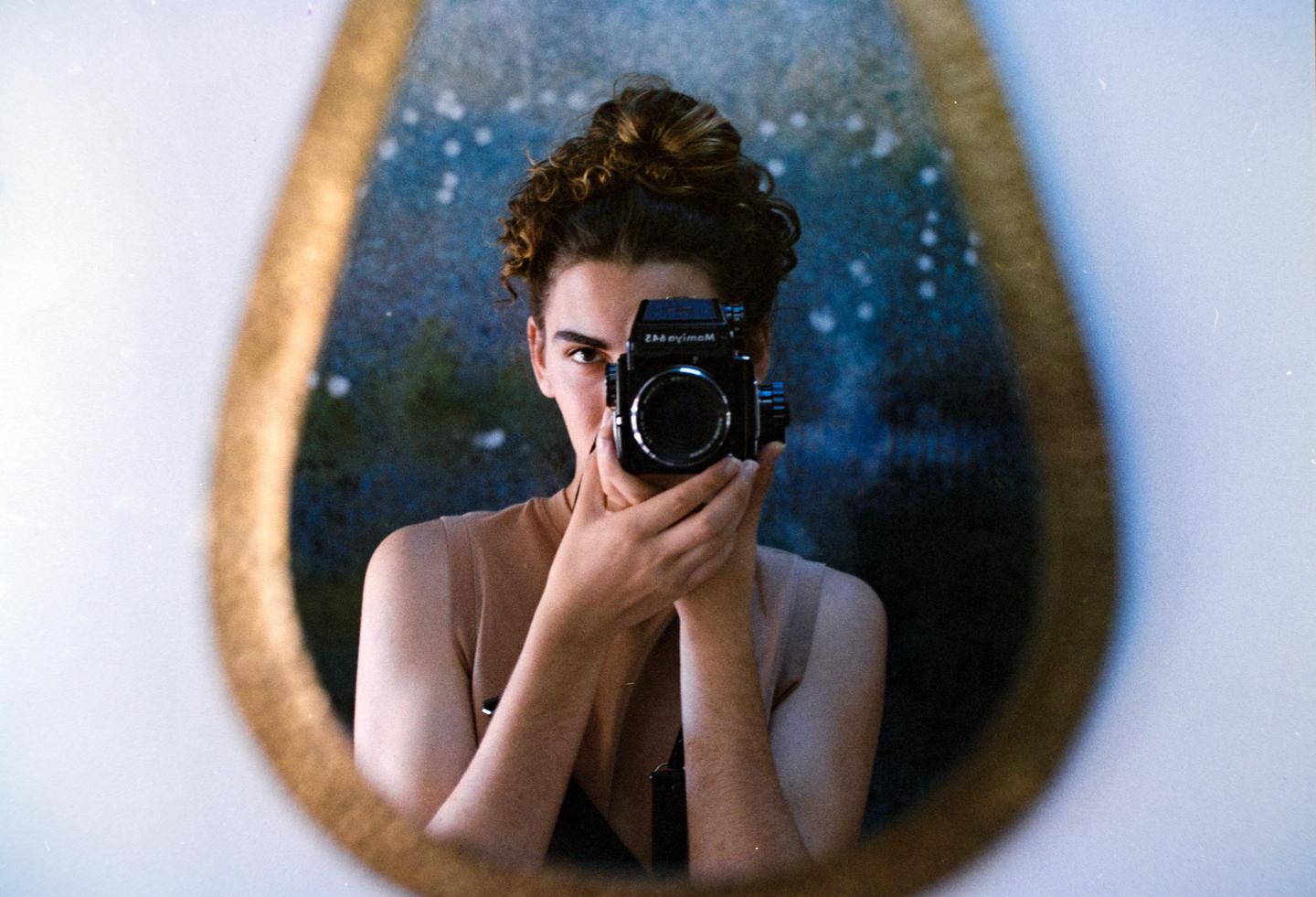 A person with curly hair in a bun holds a camera up to their face, reflected in an oval mirror.