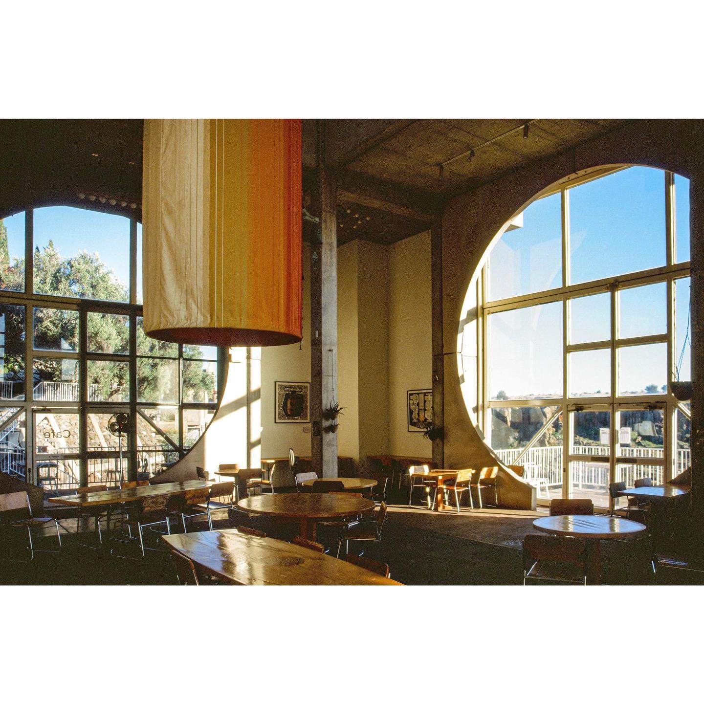 A cafe interior features large windows, wooden tables, and a cylindrical light fixture.