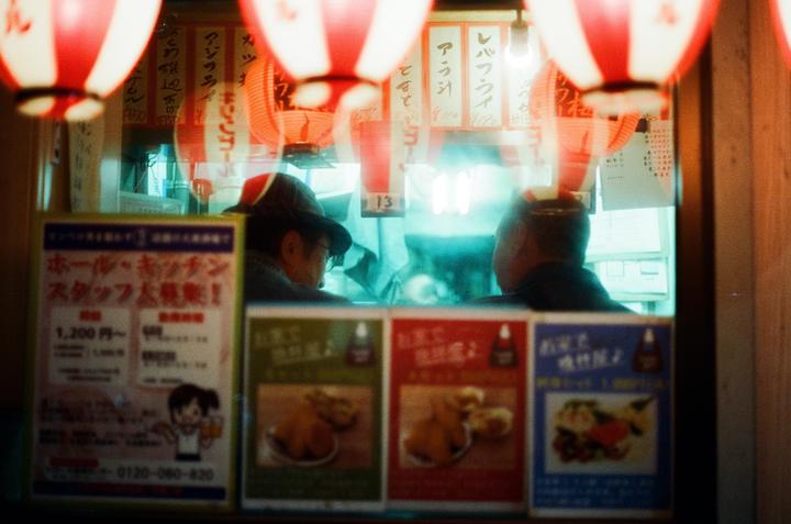 Two men are visible through a window with red and white lanterns hanging above them and advertisements below.