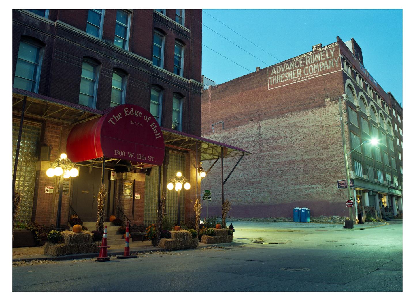 A red awning with the words "The Edge of Hell" is visible on a brick building decorated with hay bales and pumpkins.