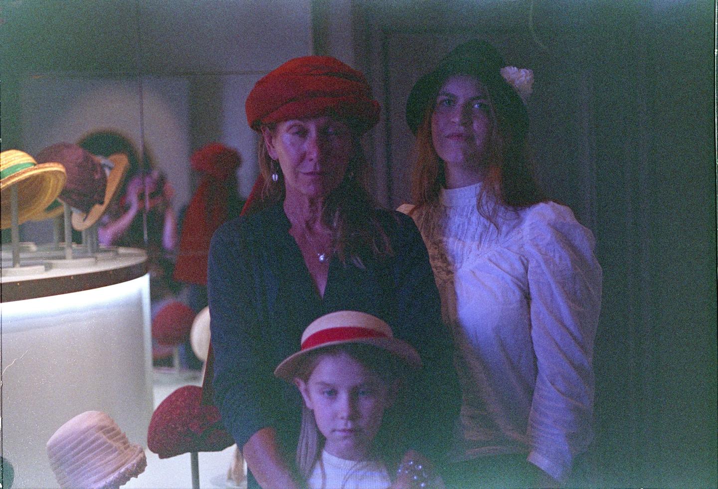 A woman in a red turban, a woman in a black hat, and a child in a straw hat pose in a room with other hats on display.