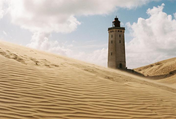 A tall lighthouse stands among sand dunes under a cloudy sky.