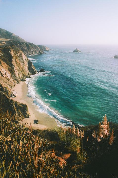 A high-angle landscape shows a sandy beach with waves crashing against a rocky coastline and a vast ocean under a clear sky.