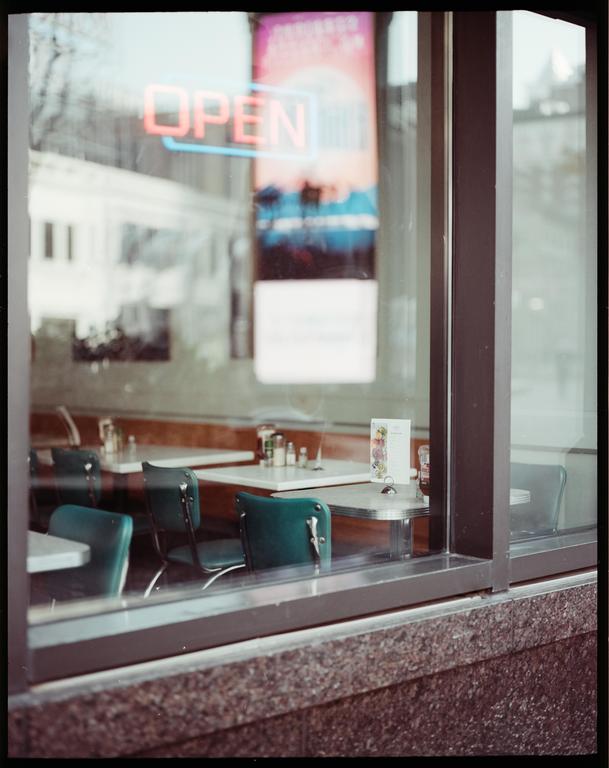 A neon "OPEN" sign is visible through the window of a diner with green chairs and white tables.