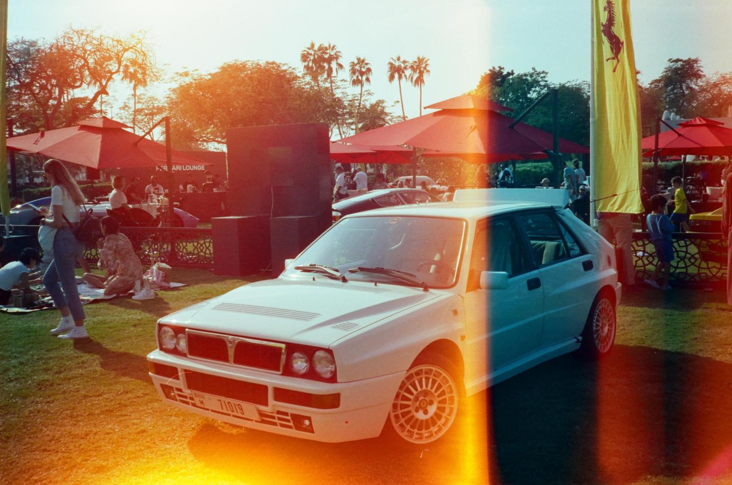 A white car is parked on a grassy area with people and red umbrellas in the background.
