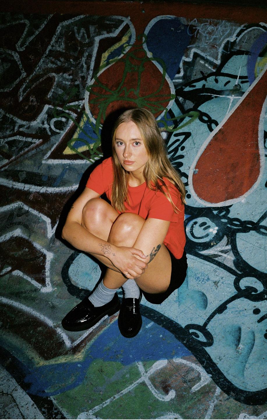 A woman in a red shirt and black shorts crouches on a graffiti-covered surface.