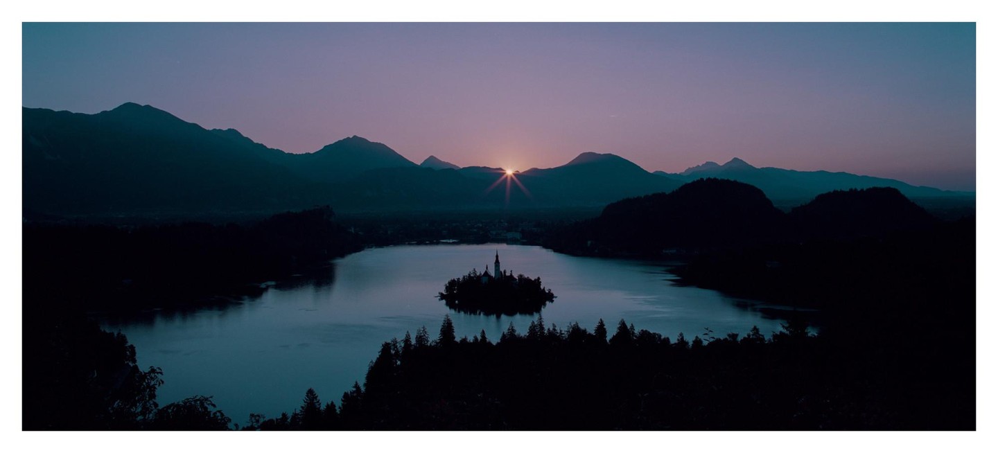 The sun rises over a lake with a small island and church, surrounded by mountains and forests.