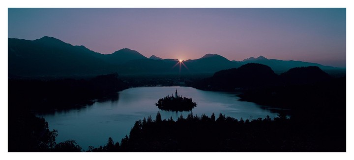 The sun rises over a lake with a small island and church, surrounded by mountains and forests.