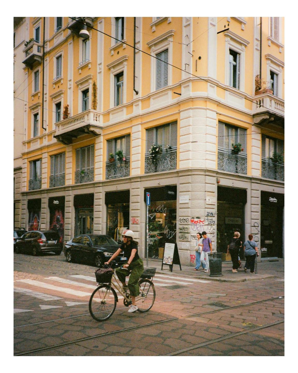 A woman rides a bicycle across a crosswalk in front of a yellow building in a city.