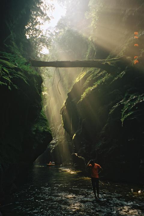 A man stands in a river in a narrow canyon with sun rays shining through the mist.
