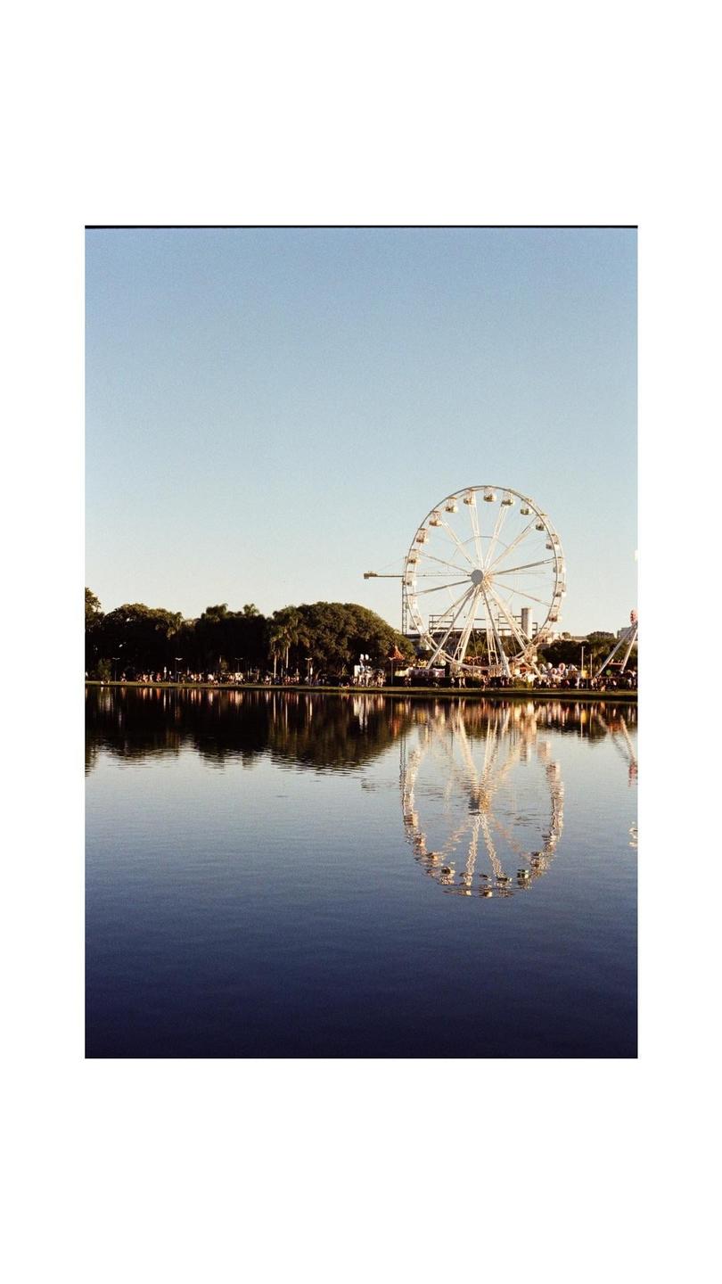 A ferris wheel is reflected in the still water of a lake with trees and people on the shore.