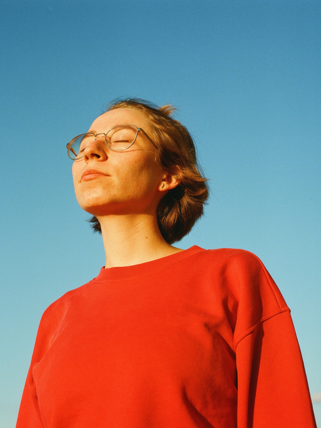 A woman with closed eyes and glasses is bathing in sunlight against a clear blue sky.
