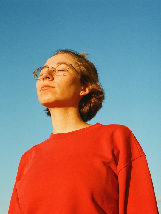 A woman with closed eyes and glasses is bathing in sunlight against a clear blue sky.