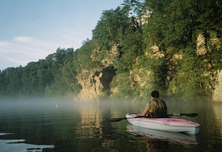 A woman in a pink kayak paddles on a misty lake surrounded by a rocky, forested shore.