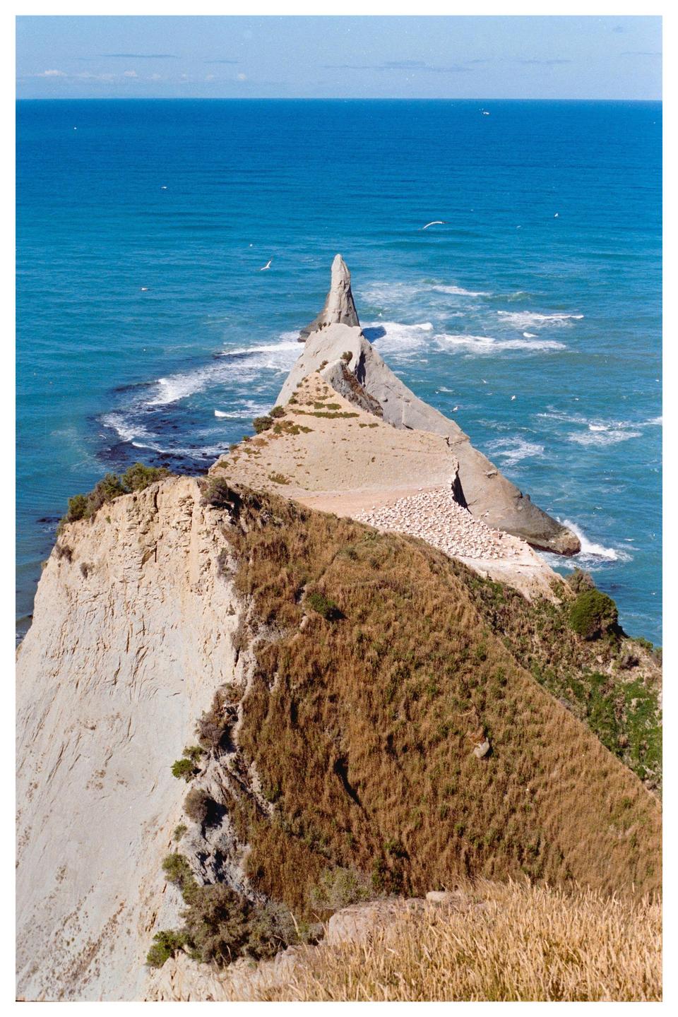 A narrow, rocky peninsula with a tall, pointed rock formation at its end extends into a blue ocean with white waves.