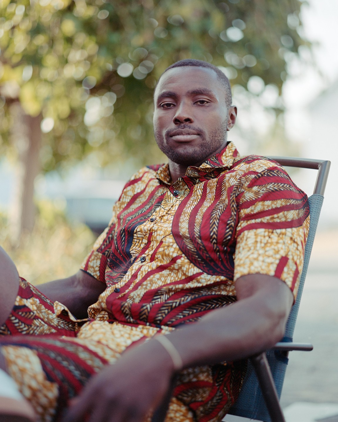 A man wearing a patterned shirt and shorts sits in a chair outdoors.