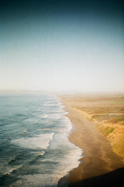 An aerial view shows waves breaking on a long, sandy beach next to a cliff.