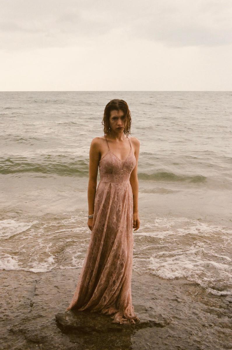 A woman in a long pink dress stands in the shallow water of a beach with waves breaking around her feet.
