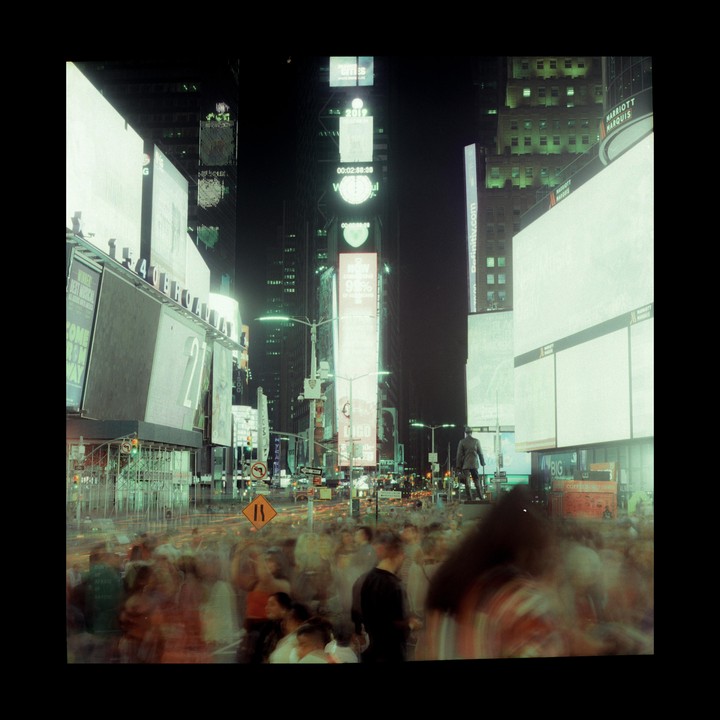 A long exposure captures the movement of people and traffic at night in Times Square, surrounded by bright billboards and buildings.