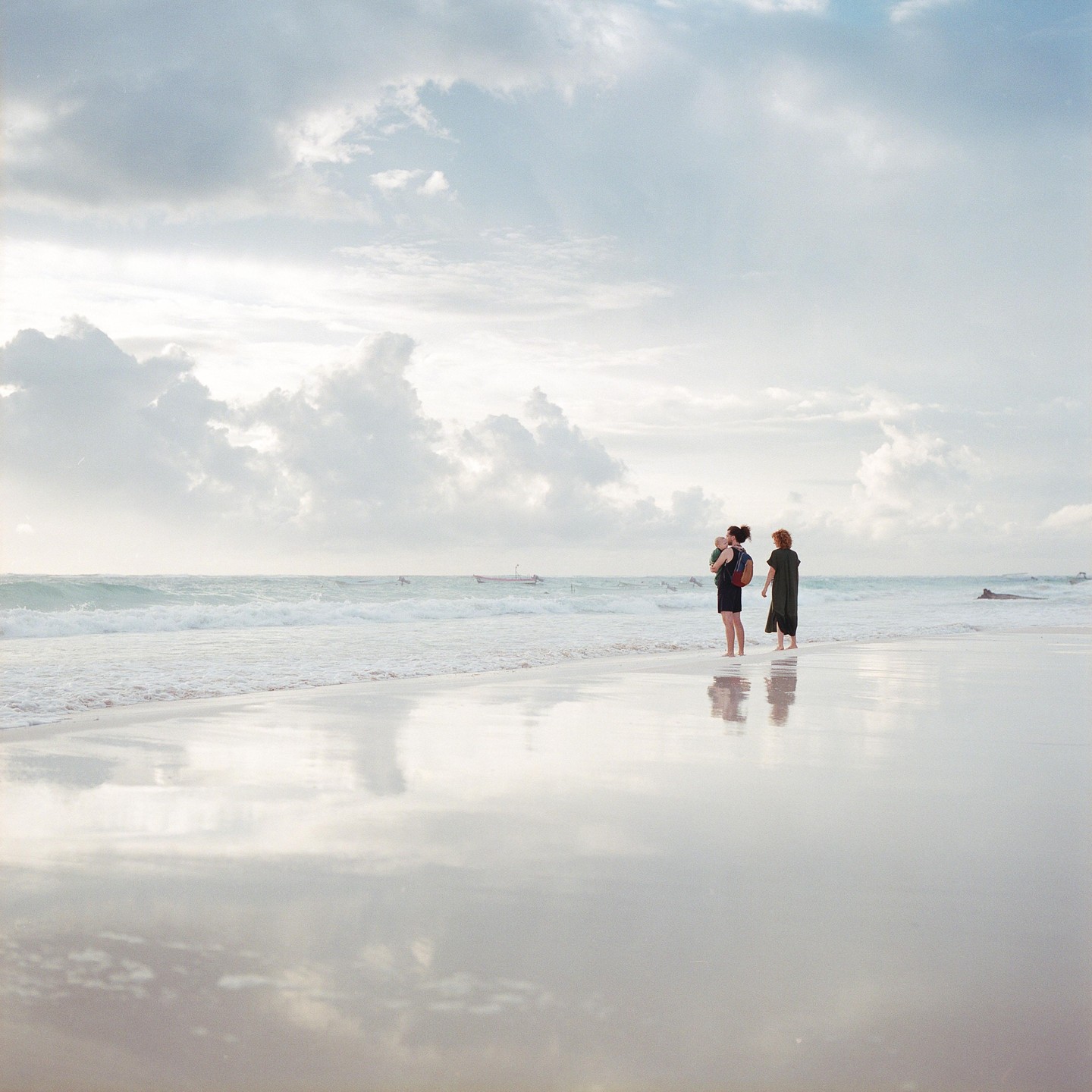 Two women stand on a wet beach with a baby in a carrier, looking out at the ocean under a cloudy sky.