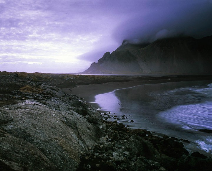 A dark mountain range looms over a beach with purple-tinged water and a rocky shore under a cloudy sky.