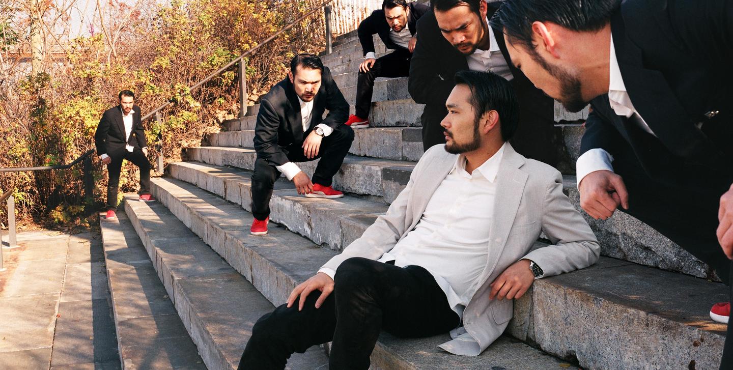 A man in a light suit sits on concrete steps surrounded by multiple versions of himself in dark suits.