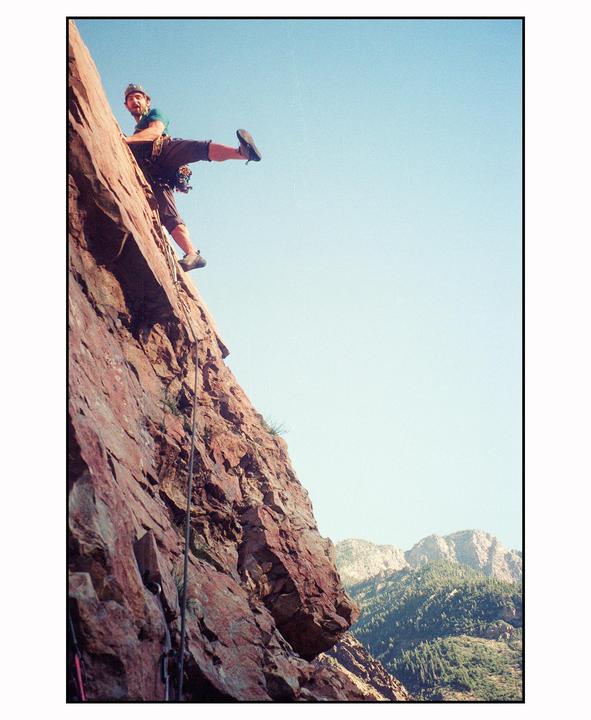 A man is rock climbing on a steep rock face with mountains in the background.