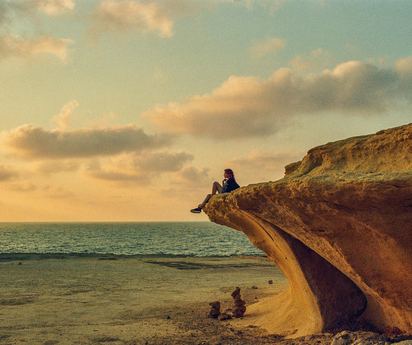 A woman sits on a rock formation overlooking the ocean at sunset.