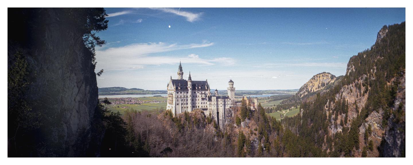 A panoramic view shows a large castle on a hill surrounded by mountains, a valley, and a body of water under a blue sky.