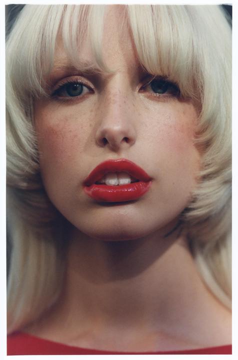 A woman with blonde hair, red lipstick, and freckles looks directly at the viewer.