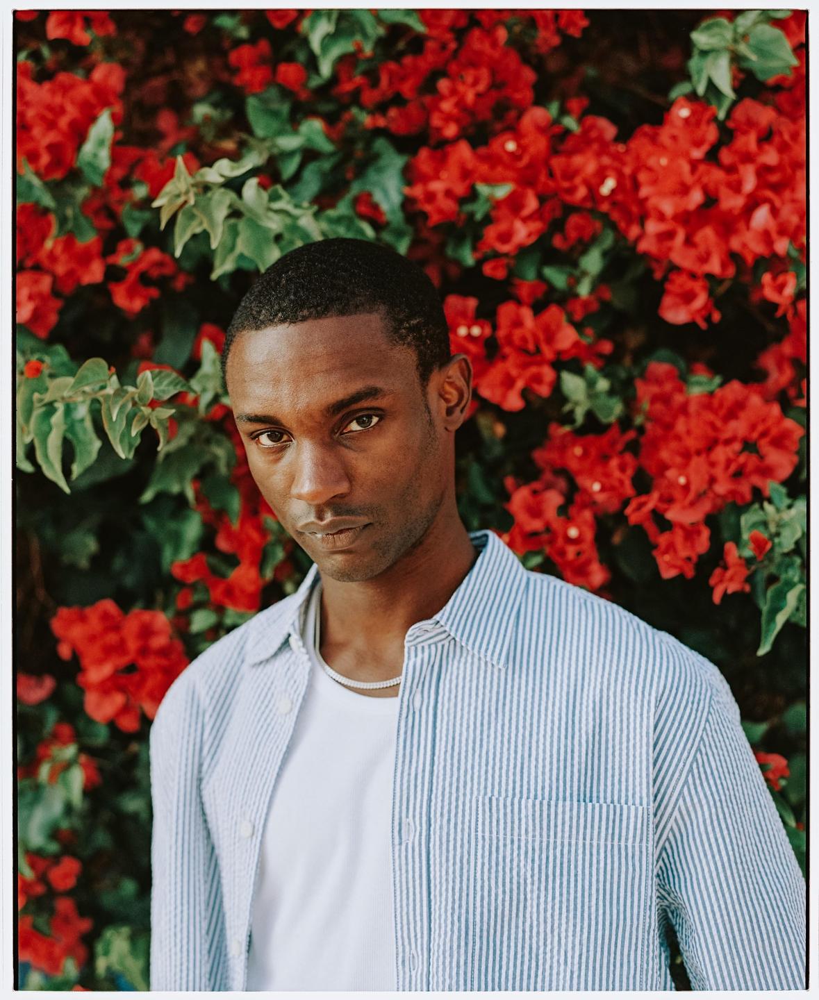 A man wearing a striped shirt and a necklace stands in front of a wall of red flowers.
