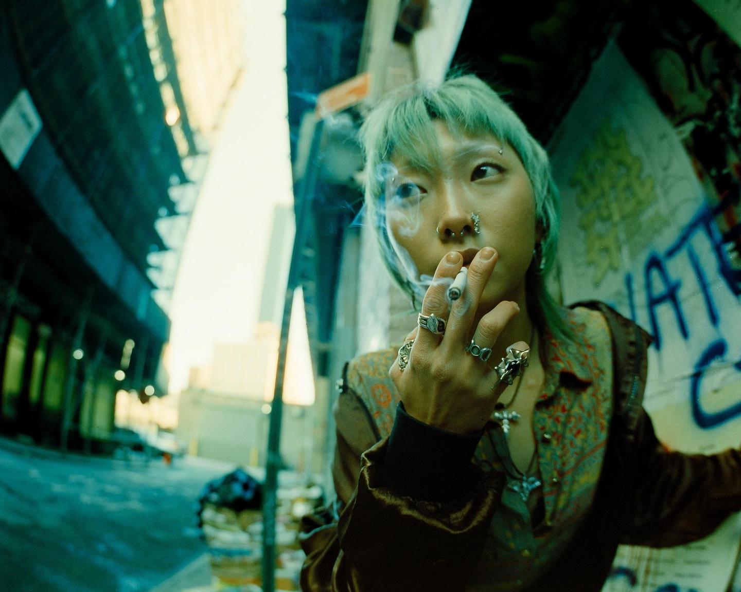 A person with green hair and multiple piercings smokes a cigarette in an alleyway with graffiti.