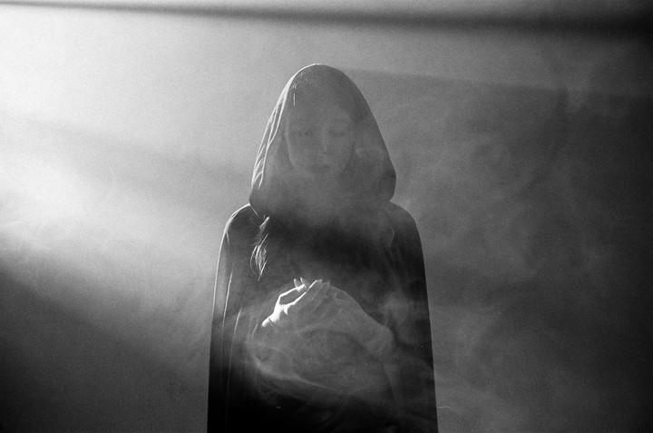 A person in a hooded cloak stands in a hazy, backlit environment, holding their hands together.