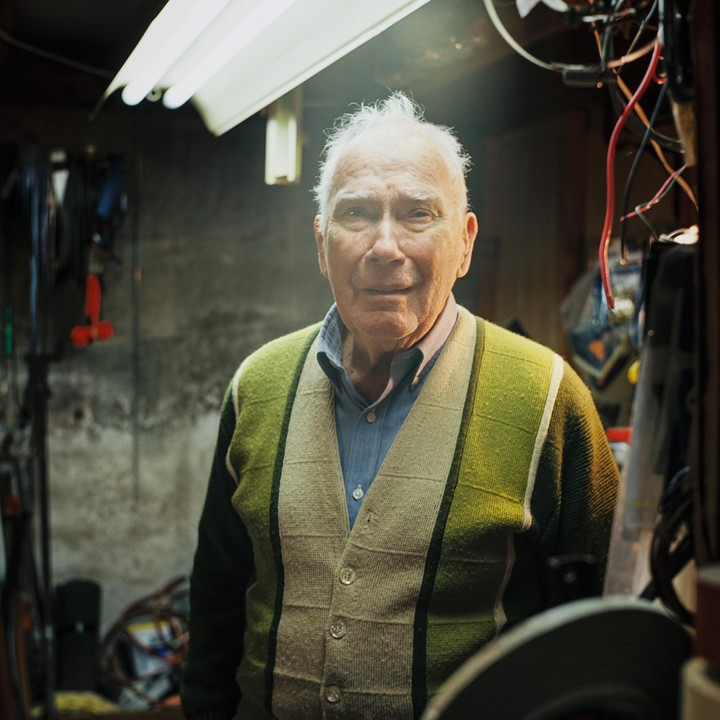An elderly man wearing a green and beige cardigan stands in a dimly lit room with exposed wires and a fluorescent light.