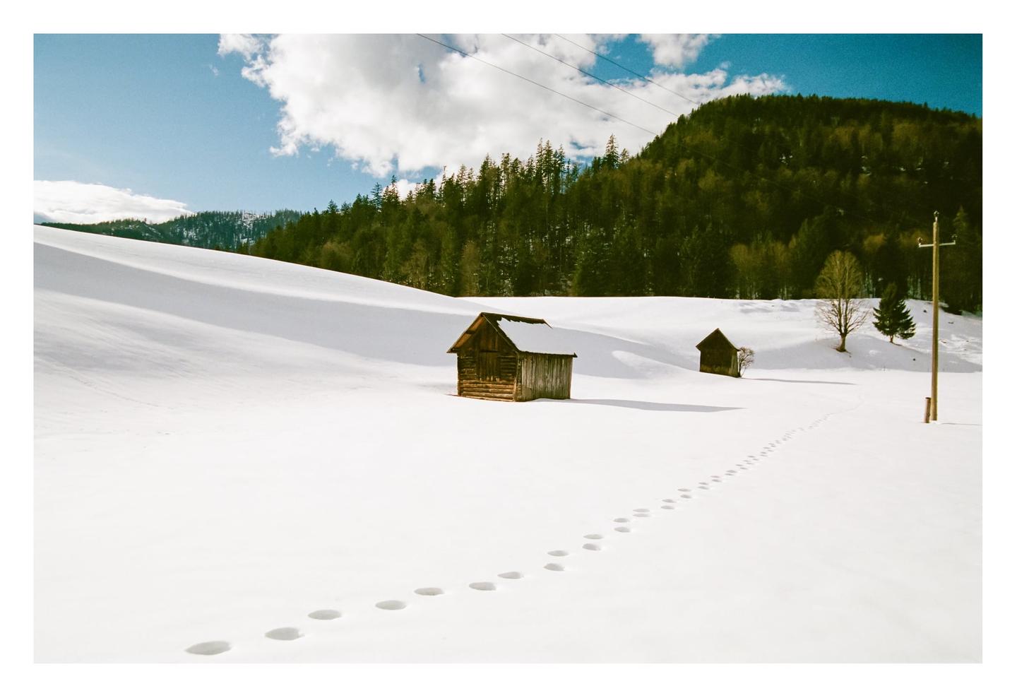 Footprints in the snow lead to a small wooden cabin in a winter landscape with a forest and mountains in the background.
