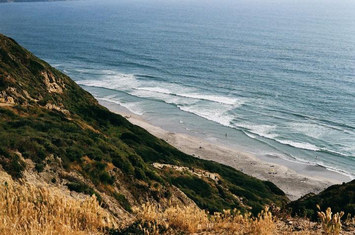 A high-angle view shows a sandy beach with ocean waves breaking on the shore next to a steep, vegetated hillside.