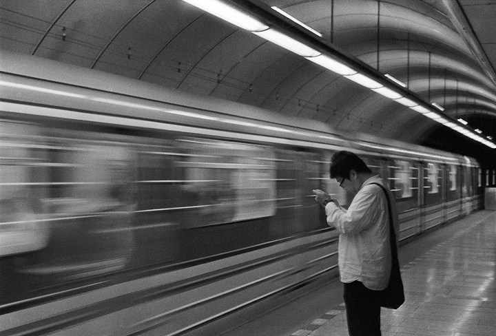 A man stands on a subway platform looking at his phone as a train speeds by.
