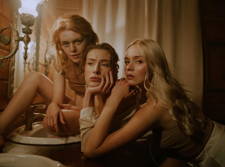 Three women pose in a dimly lit room, one sitting on a sink, another leaning on her hand, and the third reclining.