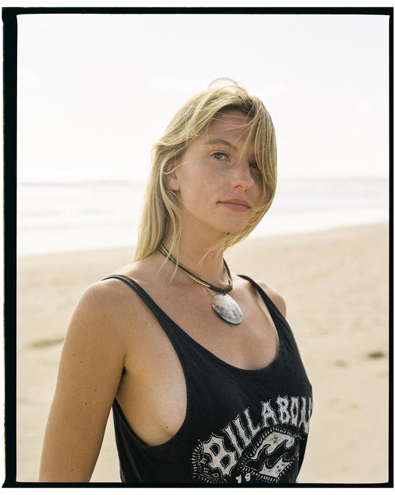 A woman with blonde hair and a necklace stands on a beach.