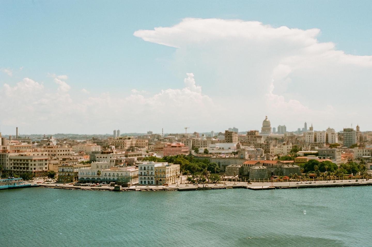 A wide shot shows the Havana cityscape with a large body of water in the foreground and a cloudy sky above.