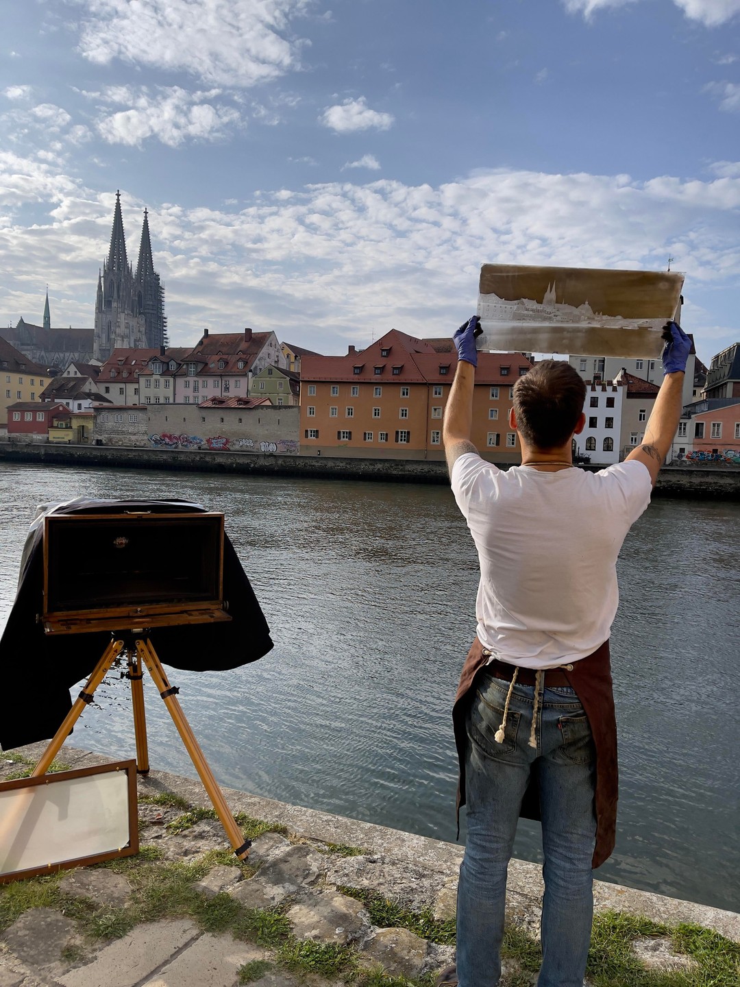 A man in an apron and gloves holds up a large negative with a cityscape on it, standing next to a river with a city and cathedral in the background.