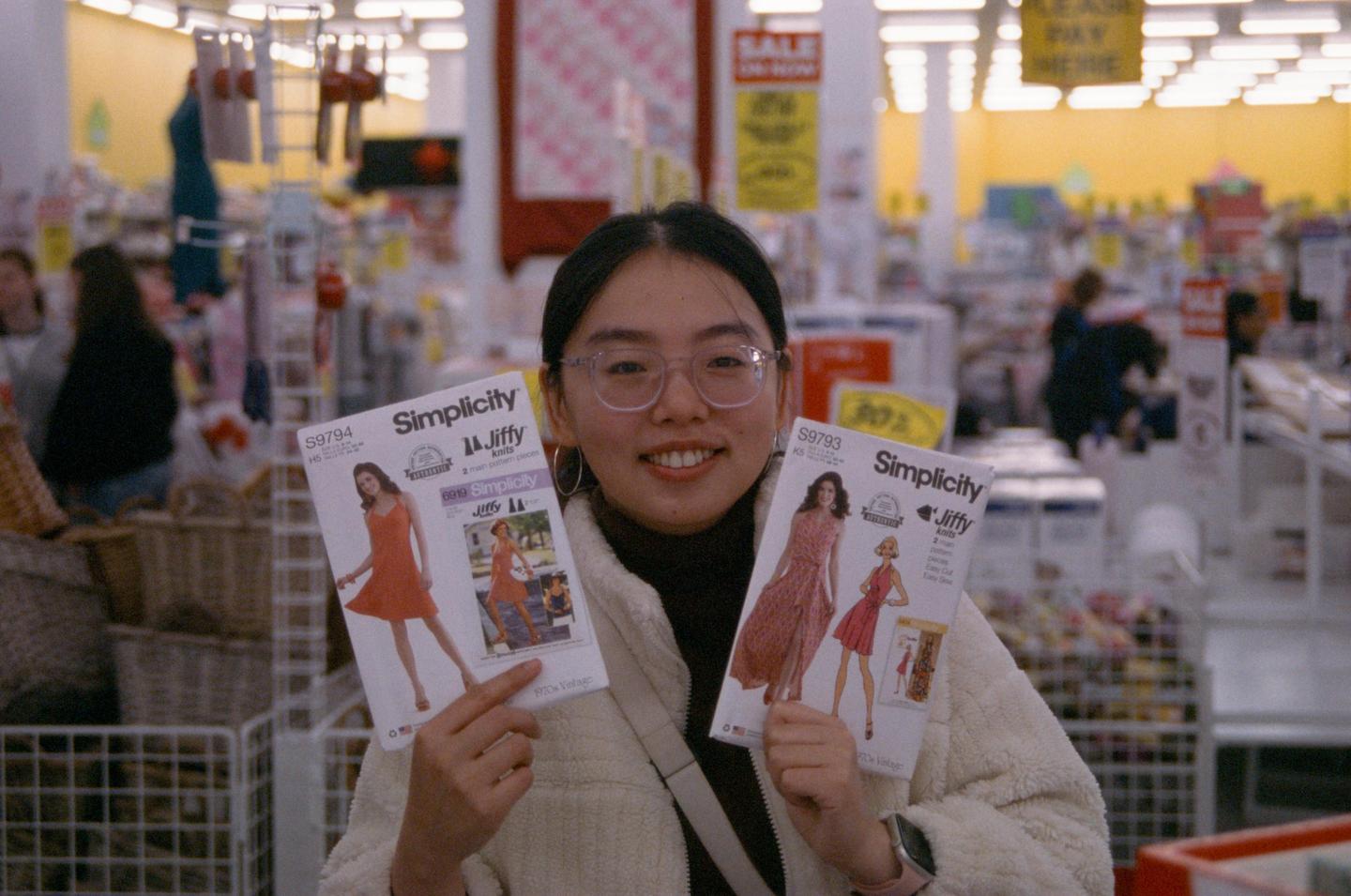 A woman in glasses smiles at the camera while holding two sewing patterns in a store.