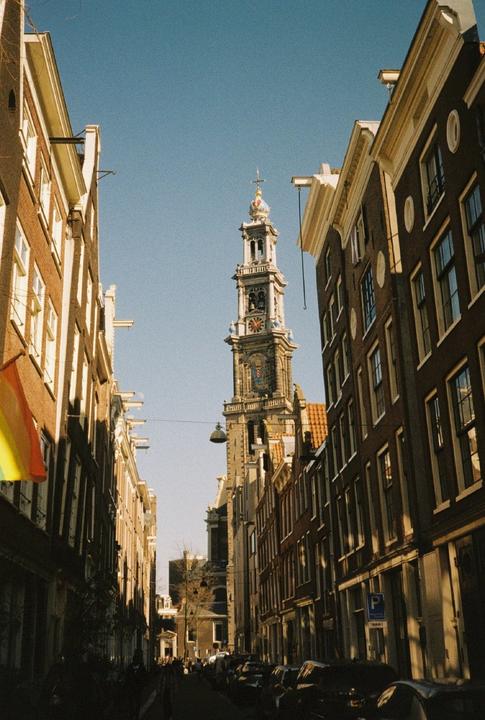 A tall church tower rises above a narrow street lined with buildings under a clear sky.