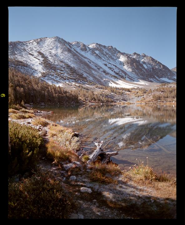A snow-capped mountain range reflects in a clear lake surrounded by forest and dry grass.