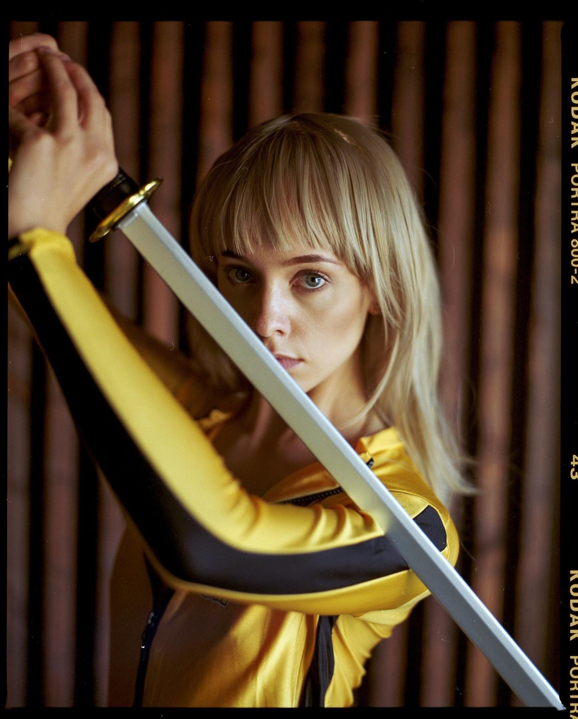 A woman in a yellow and black suit holds a sword in front of her face.