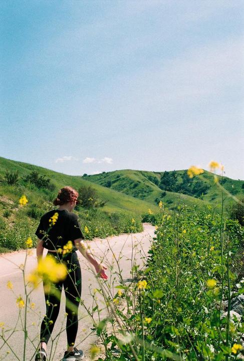 A woman walks along a winding path through green hills covered in yellow wildflowers.